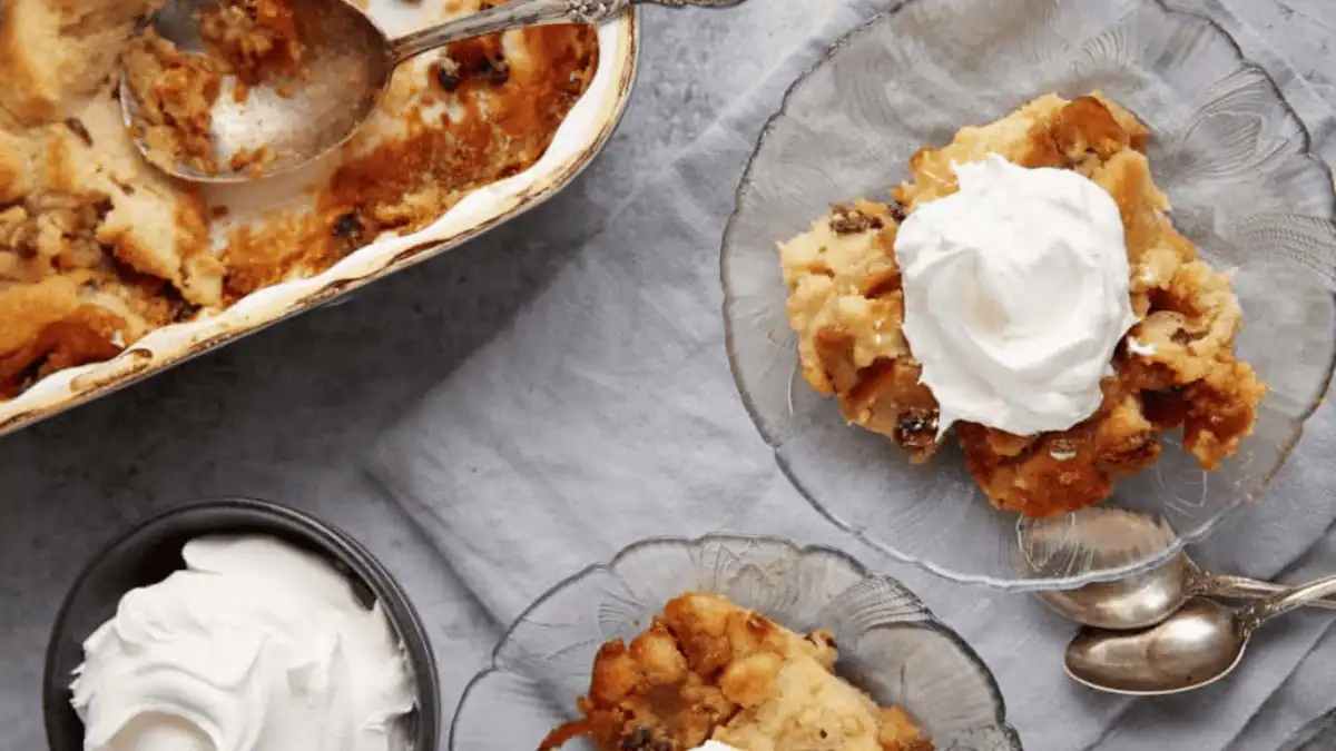 Glass plates with servings of bread pudding topped with whipped cream, a baking dish with more bread pudding, and a bowl of whipped cream on a gray surface.