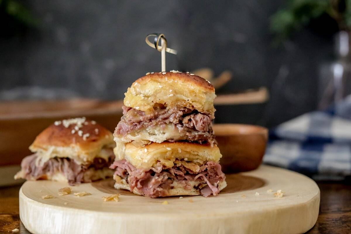 Two stacked roast beef sliders with sesame seed buns on a round wooden board, secured with a skewer; more sliders are visible in the background.