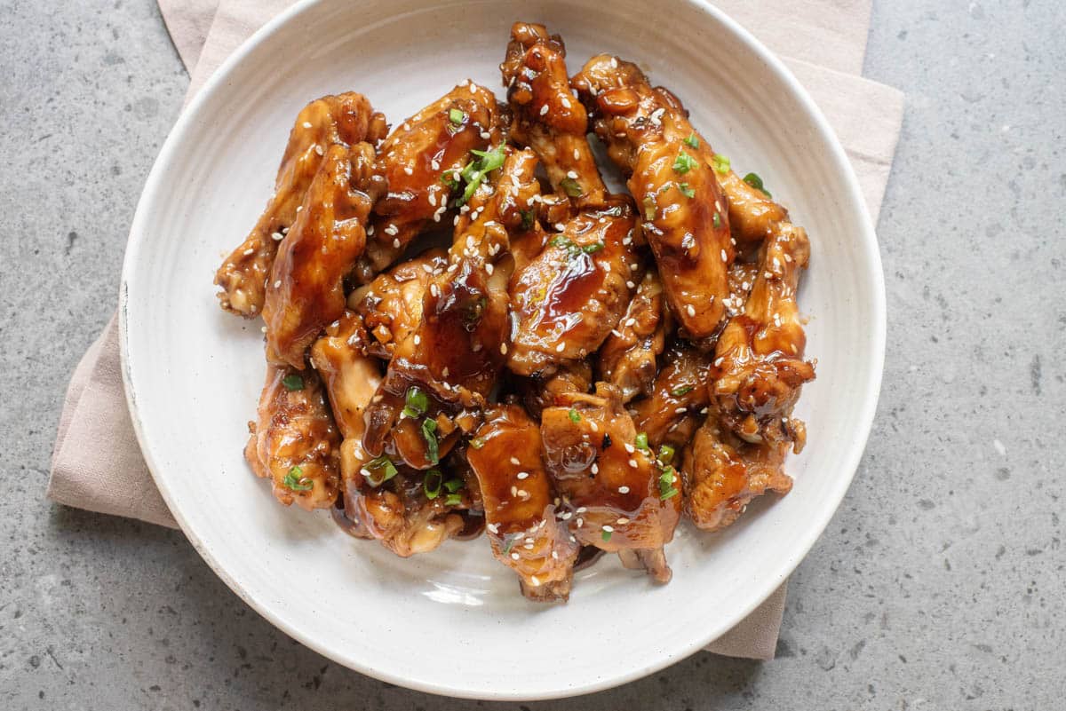 A plate of glazed chicken wings garnished with sesame seeds and chopped green onions, placed on a beige napkin atop a gray surface.