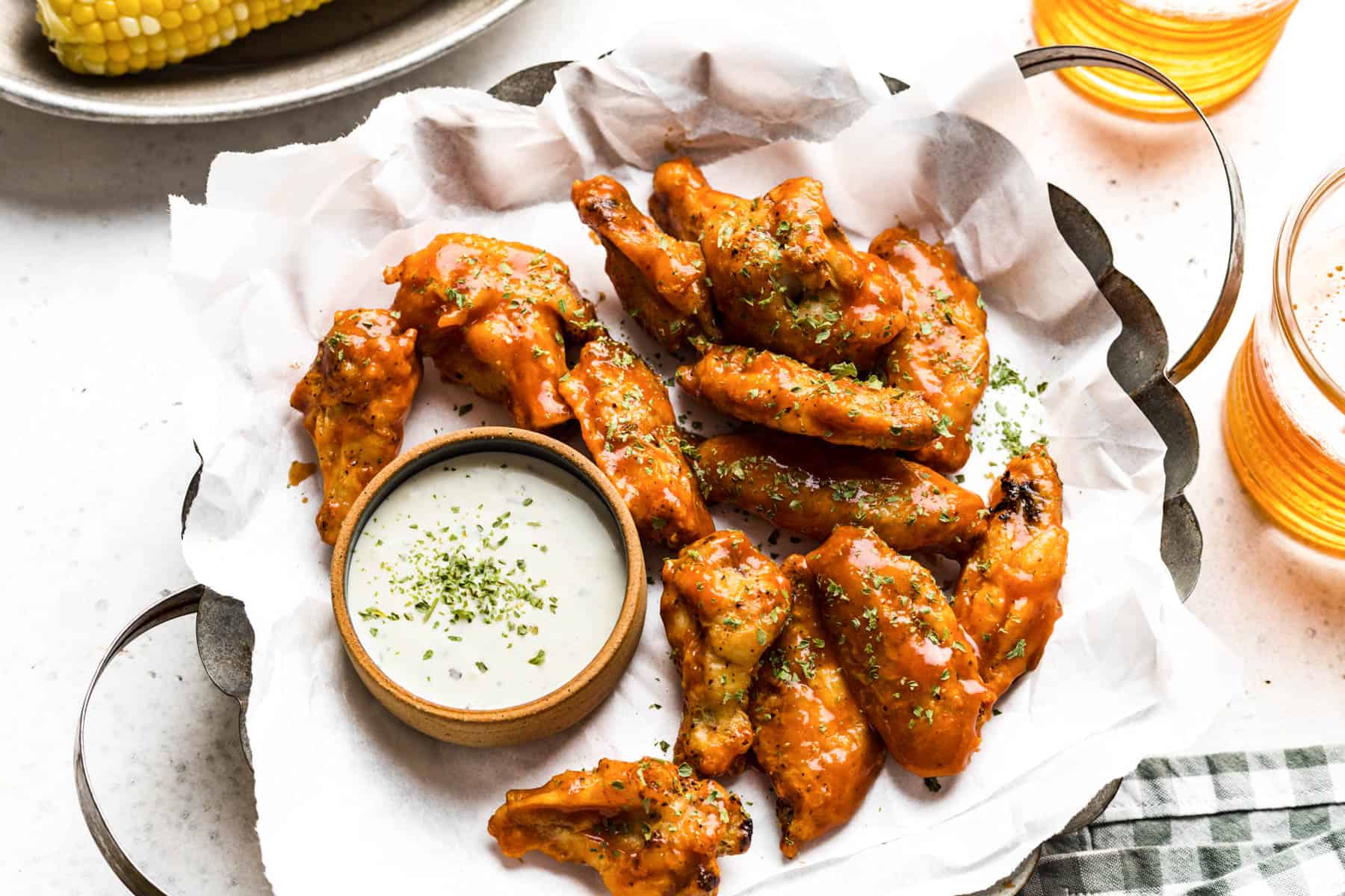 A plate of buffalo chicken wings garnished with herbs, served with a side of ranch dipping sauce on parchment paper. Corn on the cob and drinks are partially visible.