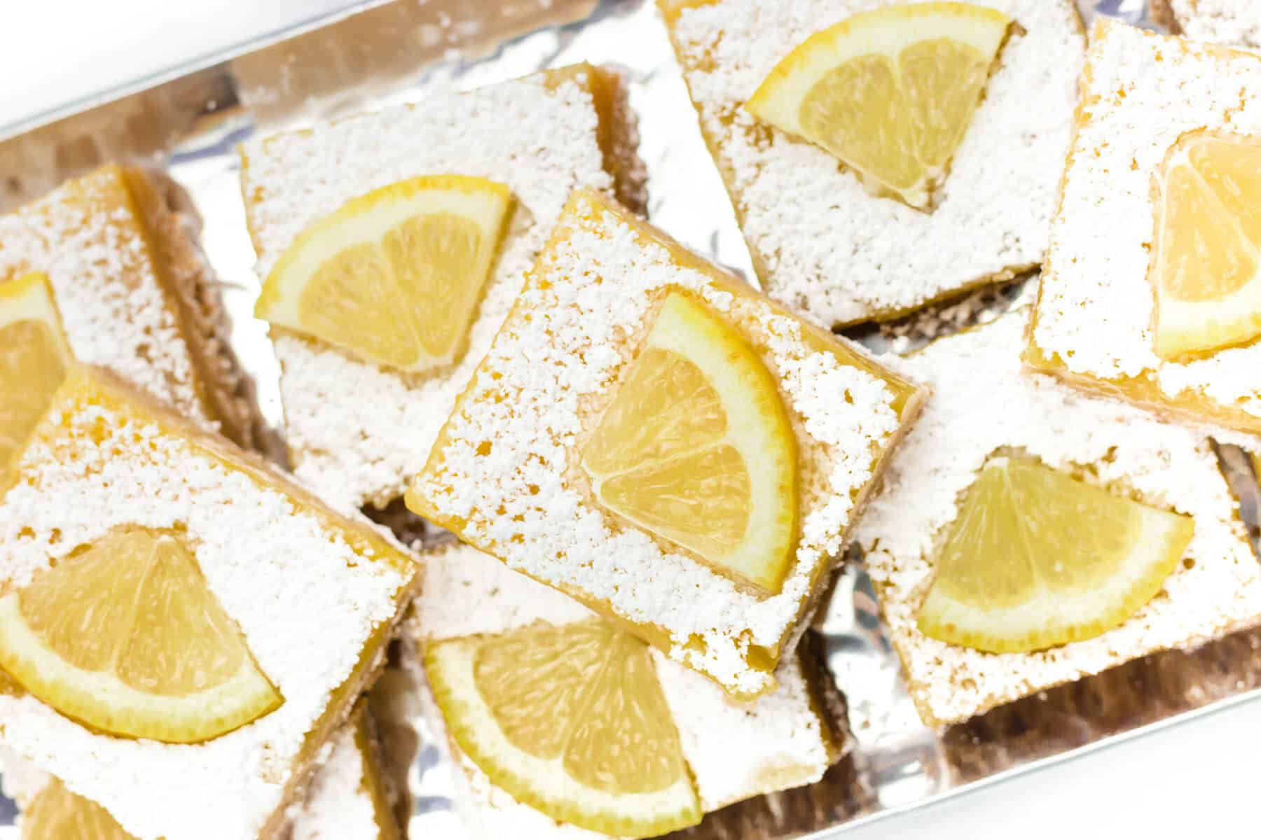 A tray of lemon bars topped with powdered sugar and a slice of lemon on each square.