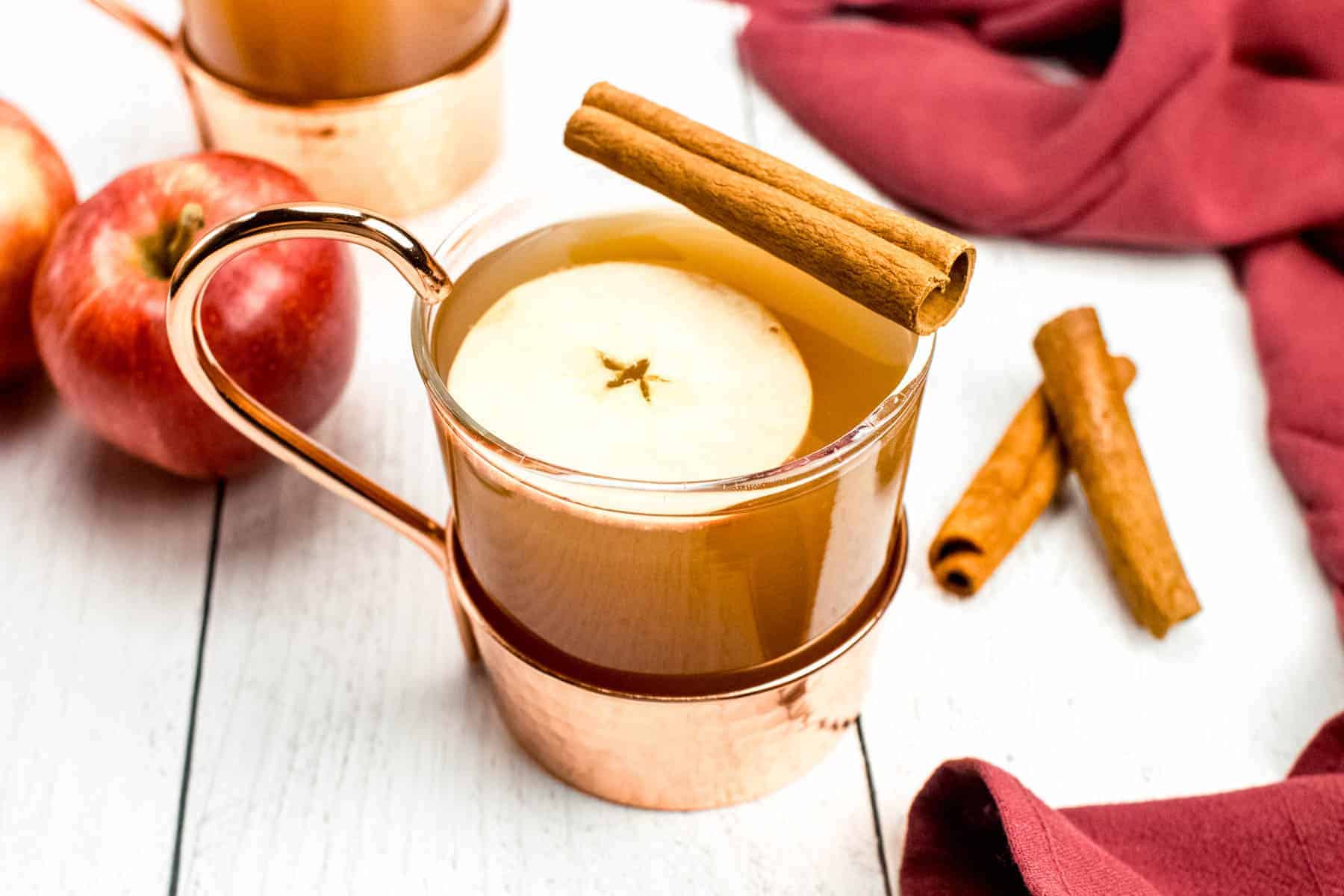 A copper mug filled with apple cider, garnished with a cinnamon stick and an apple slice, sits on a white wooden surface with apples and a red cloth nearby.