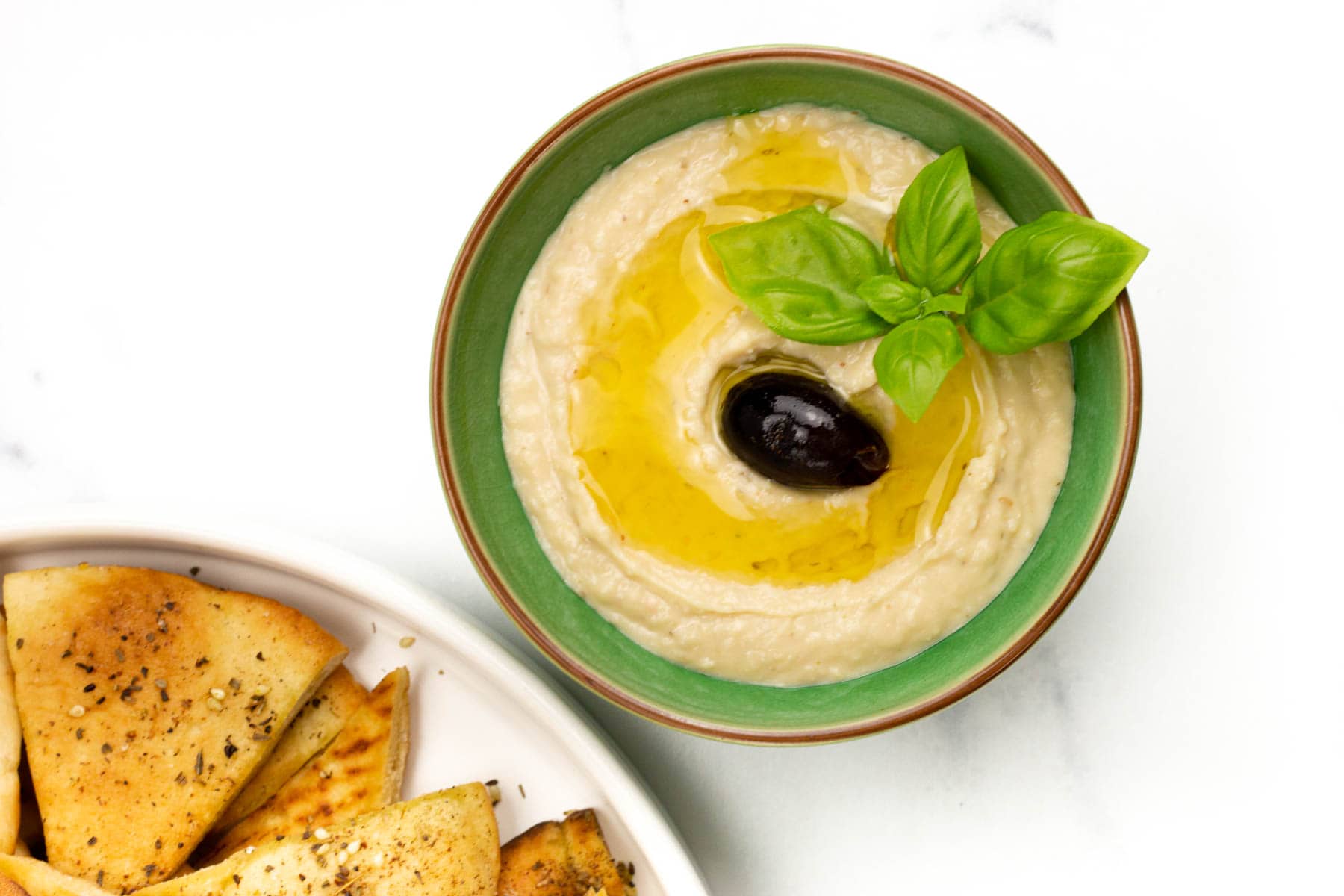 A bowl of hummus topped with olive oil, a black olive, and basil leaves, next to a plate of seasoned pita chips.