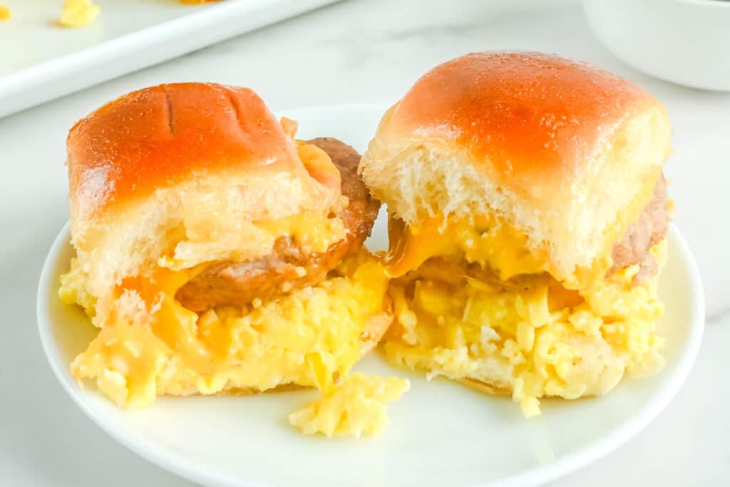 Two breakfast sliders on a plate, each with scrambled eggs, melted cheese, and sausage between soft, shiny buns.