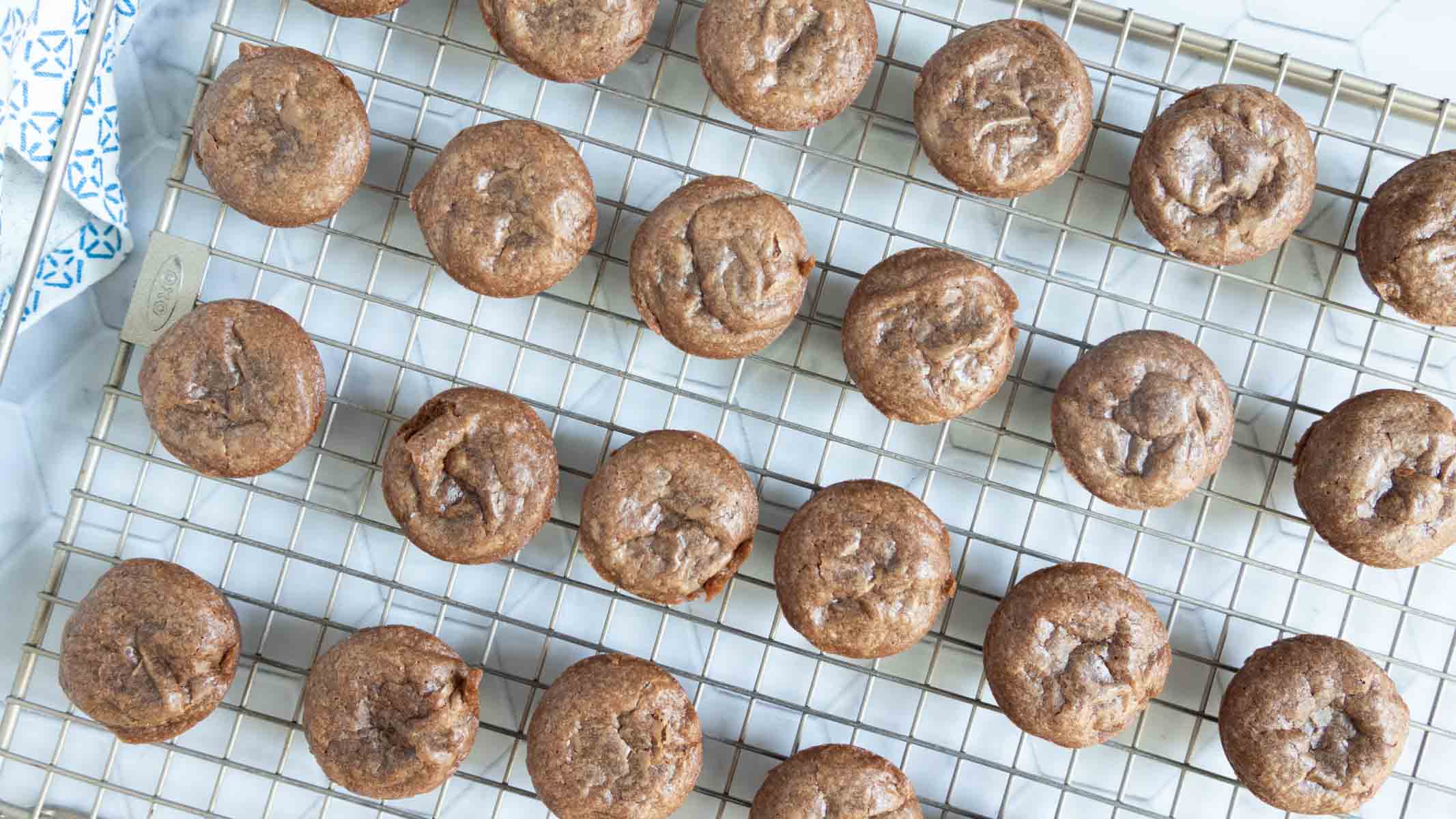 Small chocolate muffins cooling on a metal wire rack, arranged in rows on a white surface.