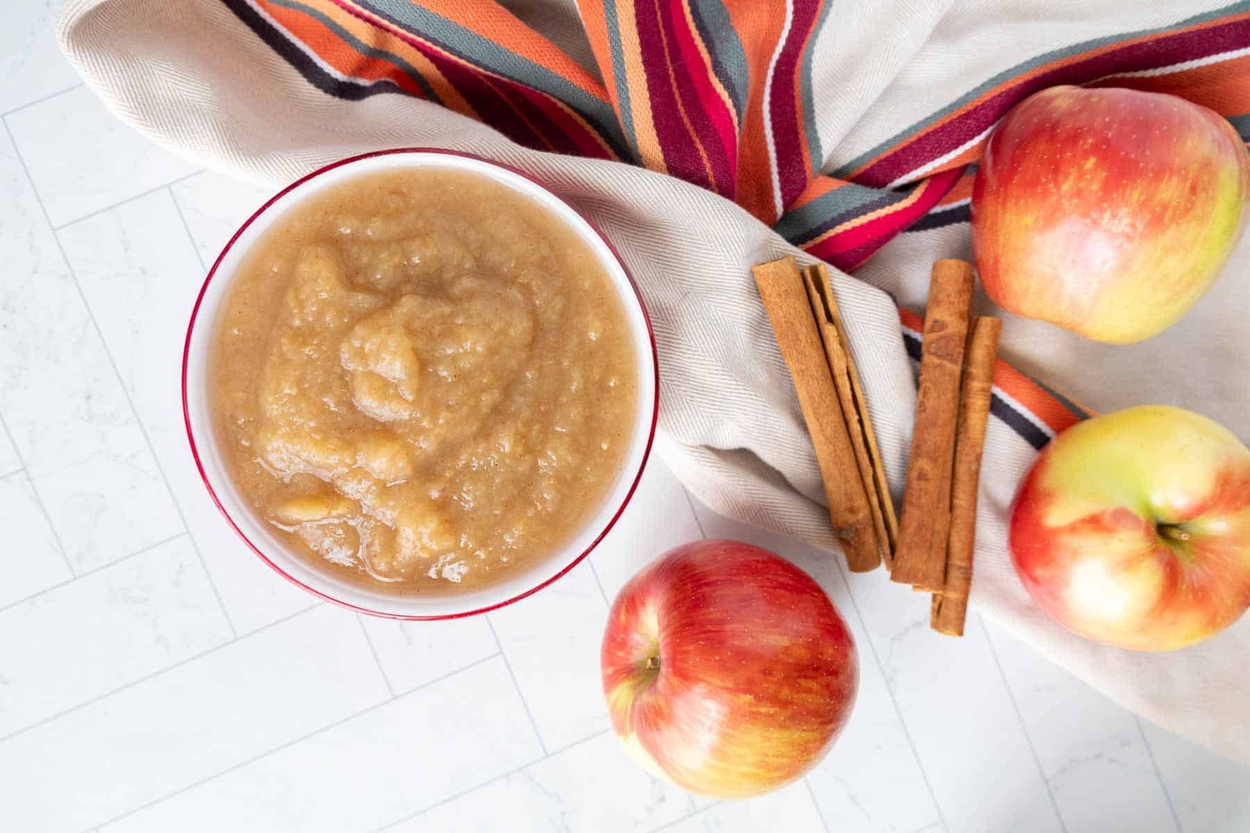 A bowl of applesauce next to two whole apples, cinnamon sticks, and a patterned cloth on a white surface.