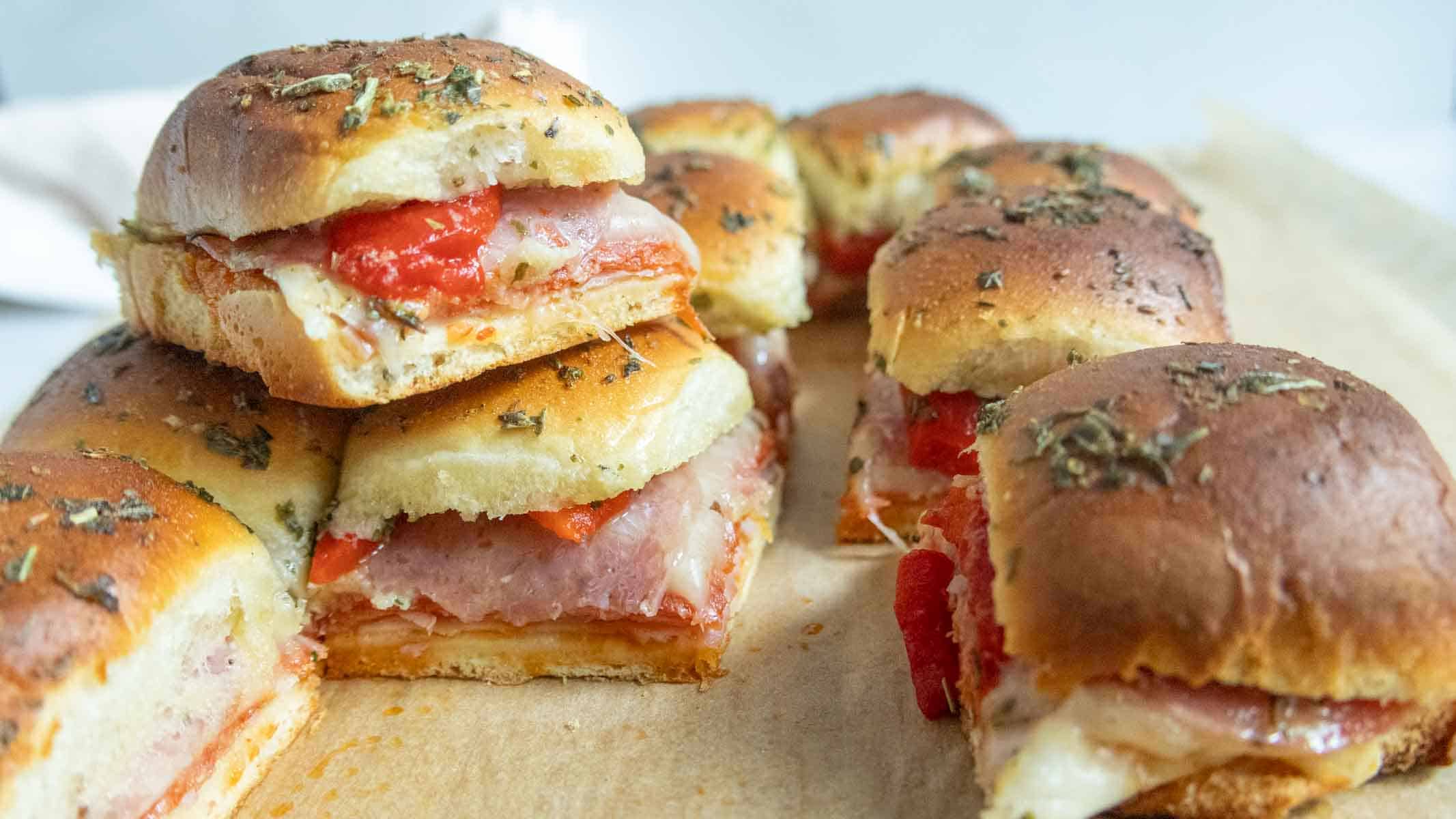 A close-up of baked sandwiches with ham, cheese, and roasted red peppers on herb-topped bread, arranged on a parchment-lined surface.