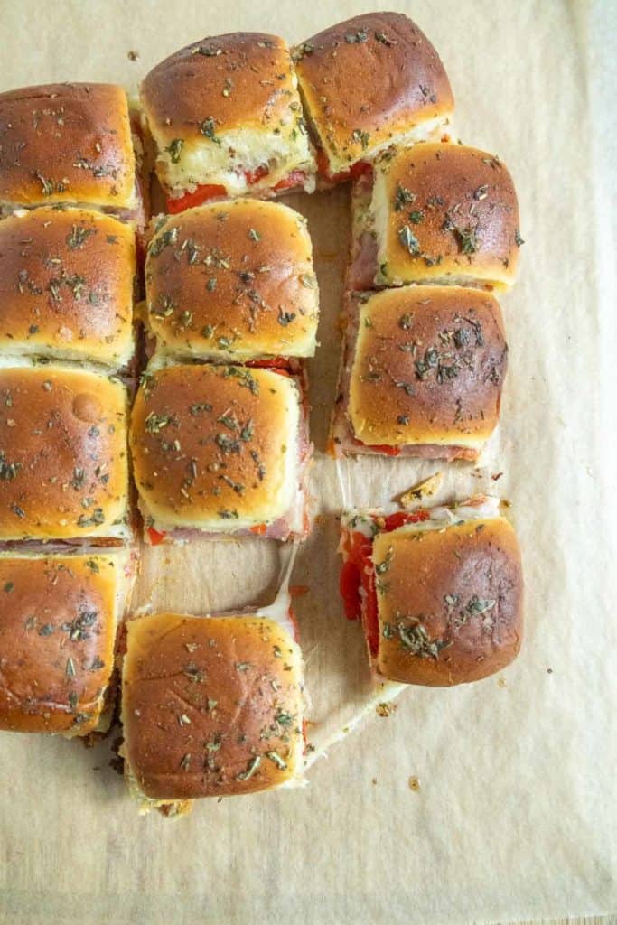 A batch of baked pull-apart slider sandwiches with melted cheese, herbs, and sliced filling on parchment paper, with one slider partially removed.