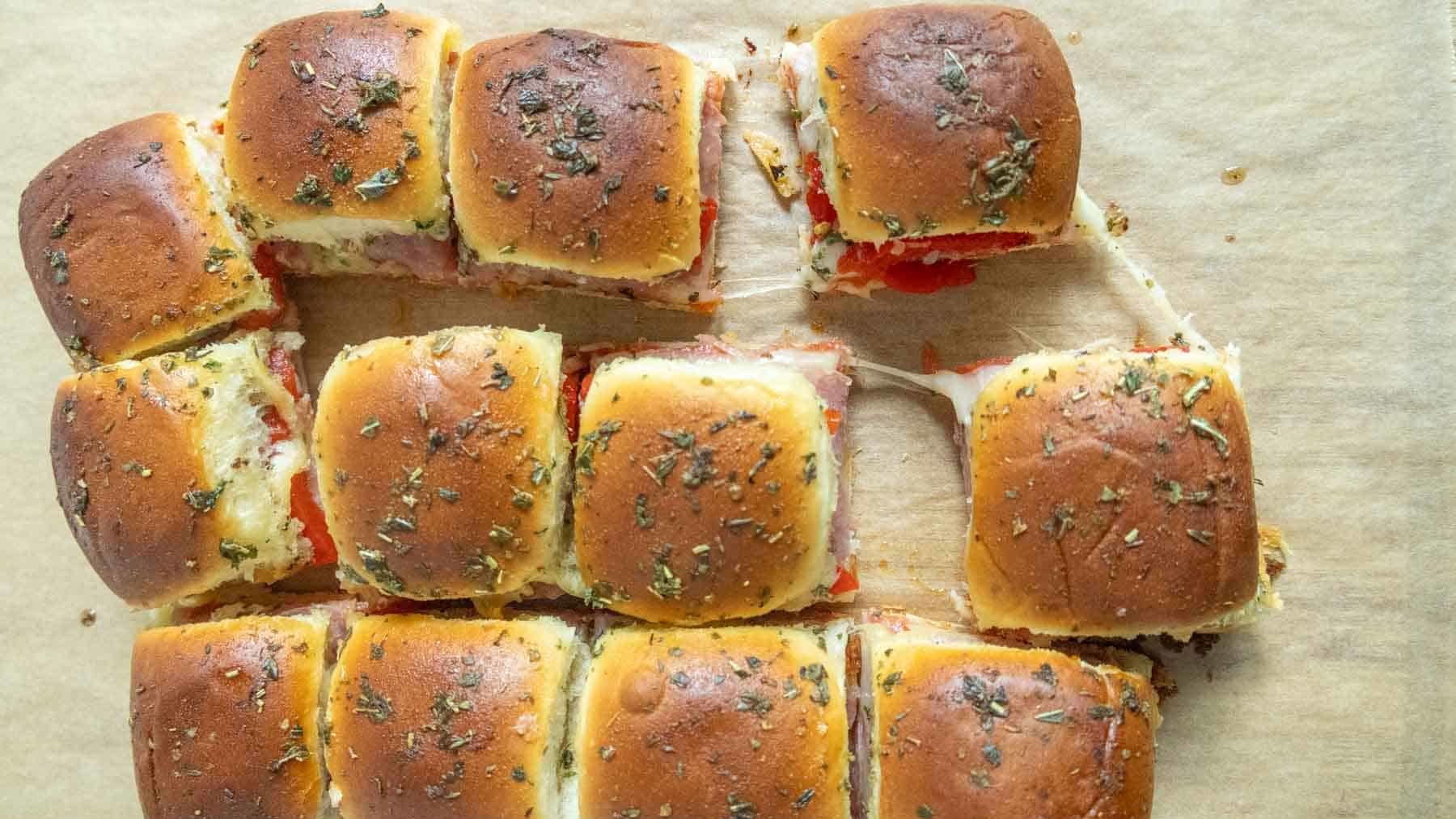 A batch of baked pull-apart slider sandwiches with melted cheese, herbs, and sliced filling on parchment paper, with one slider partially removed.