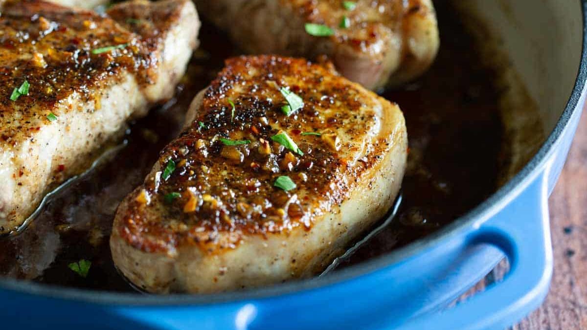 Three cooked pork chops with herbs in a blue cast iron skillet on a wooden surface.