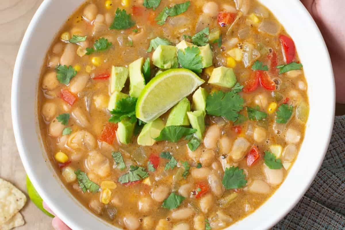 A bowl of white bean chili topped with avocado, lime wedge, and cilantro, containing corn and diced red bell peppers.