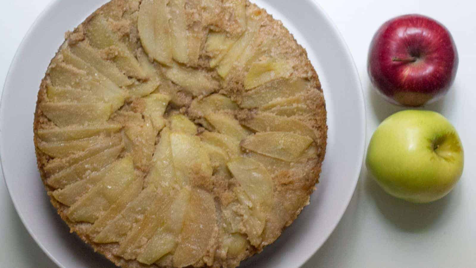 A round upside-down apple cake on a white plate next to a red apple and a green apple on a white surface.