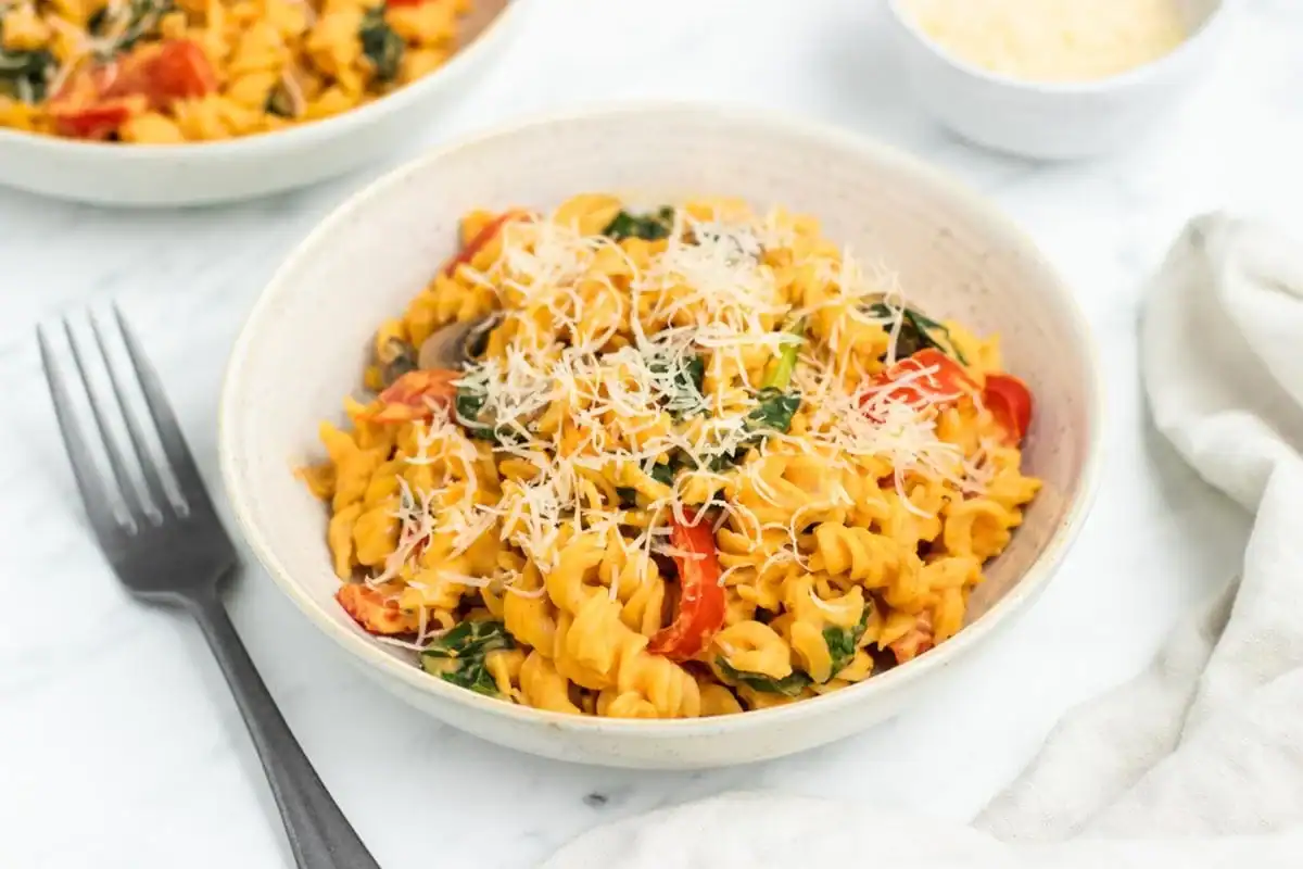 A bowl of rotini pasta mixed with vegetables and topped with grated cheese, next to a fork on a white surface.
