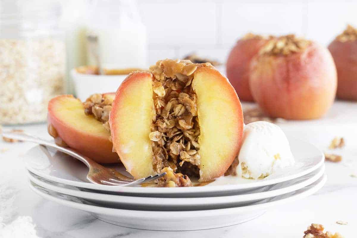 A halved baked peach filled with oat crumble is served on a plate with a scoop of vanilla ice cream and a fork.