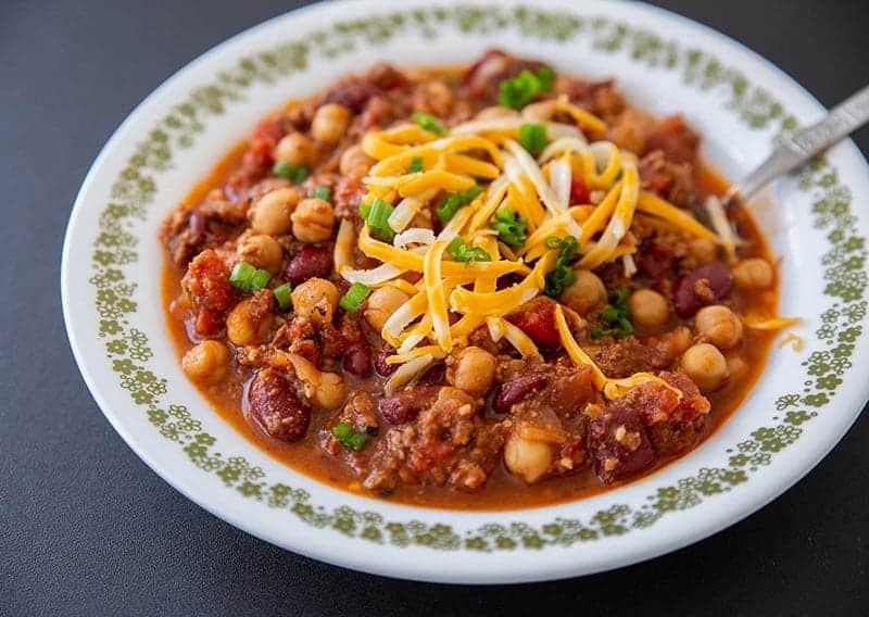 A bowl of chili with ground meat, beans, chickpeas, diced tomatoes, and shredded cheese, garnished with chopped green onions.