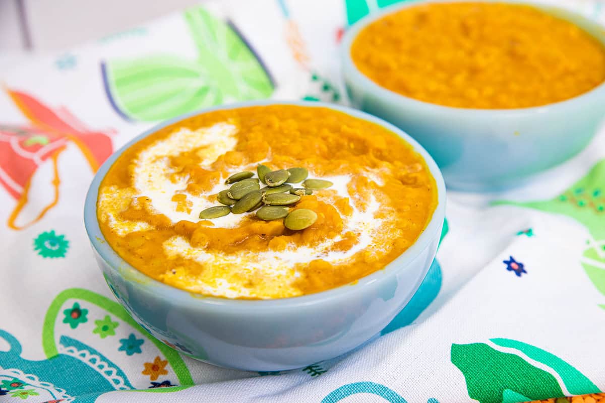 A bowl of orange soup with a swirl of cream and pumpkin seeds on top, placed on a colorful patterned cloth, with another bowl in the background.