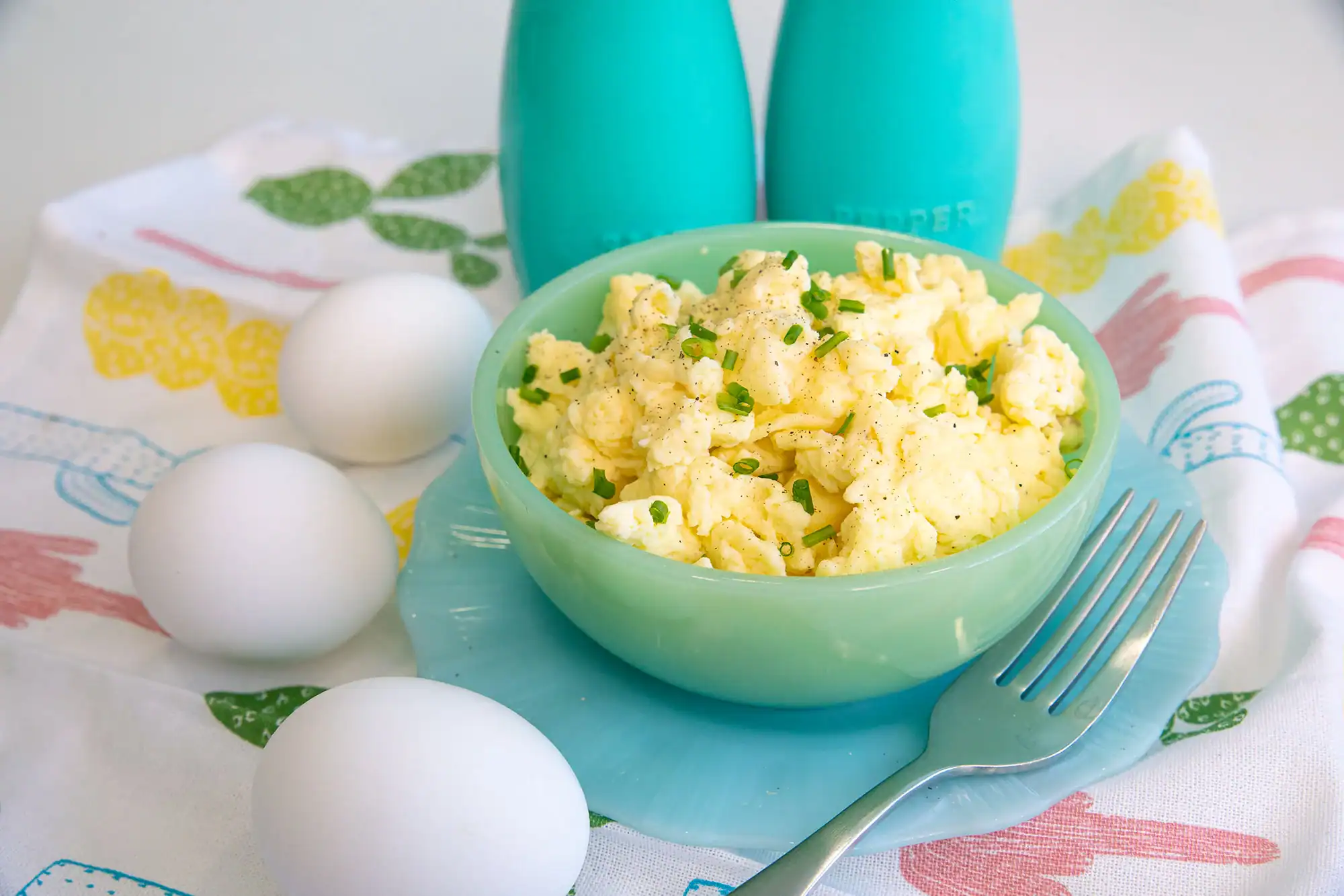 A green bowl of scrambled eggs topped with chopped chives sits on a blue plate, next to three whole eggs, a fork, and two blue salt and pepper shakers.