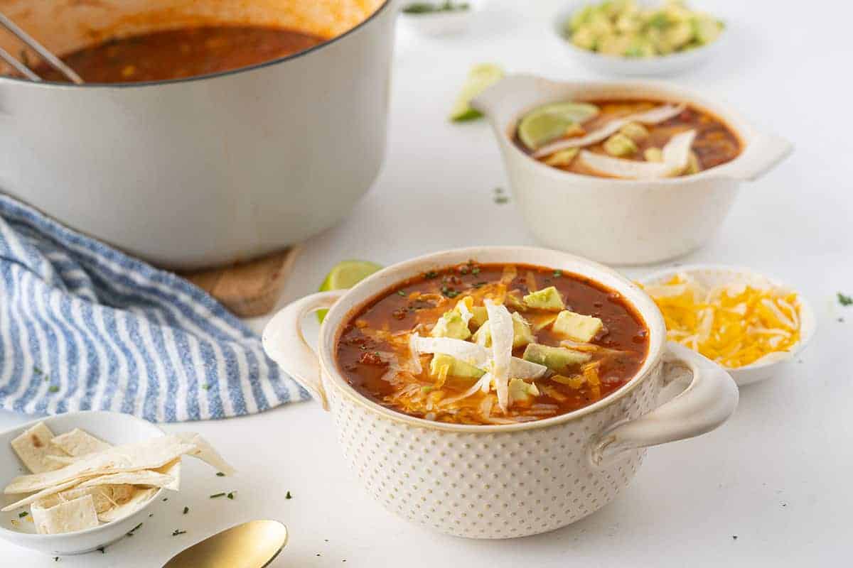 A bowl of tortilla soup topped with avocado, shredded cheese, and tortilla strips sits on a table next to a pot, another bowl, and small dishes with garnishes.