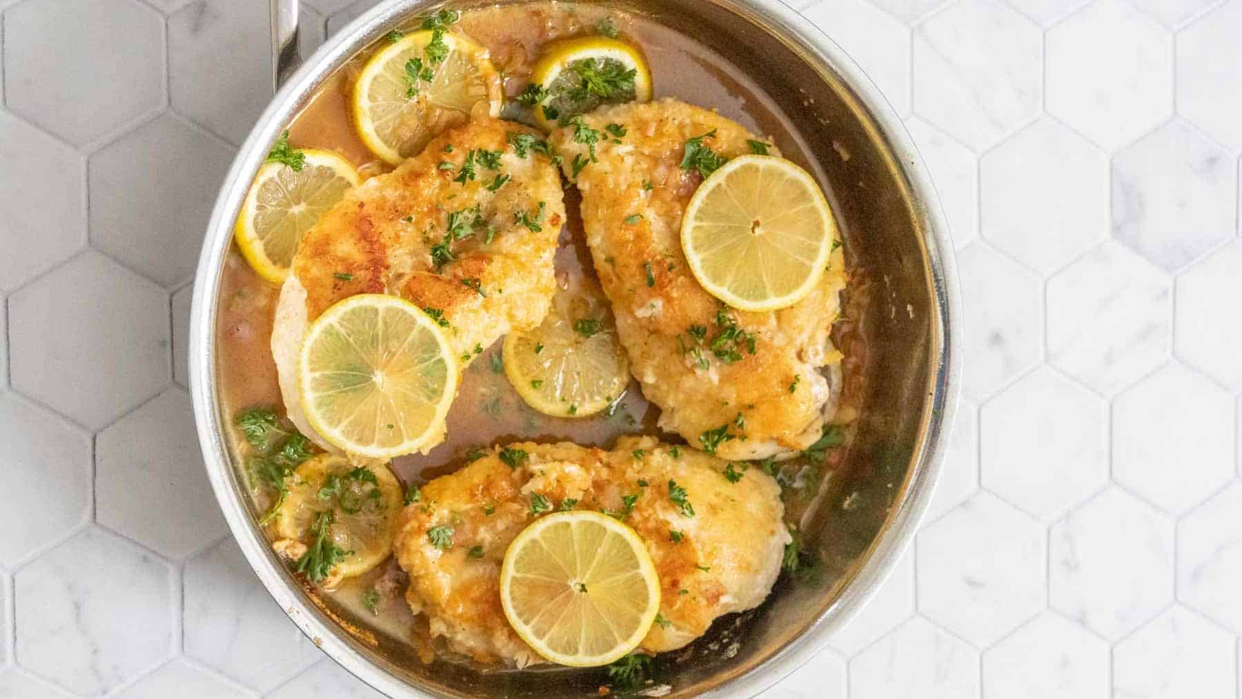 Skillet with cooked chicken breasts, lemon, and parsley.