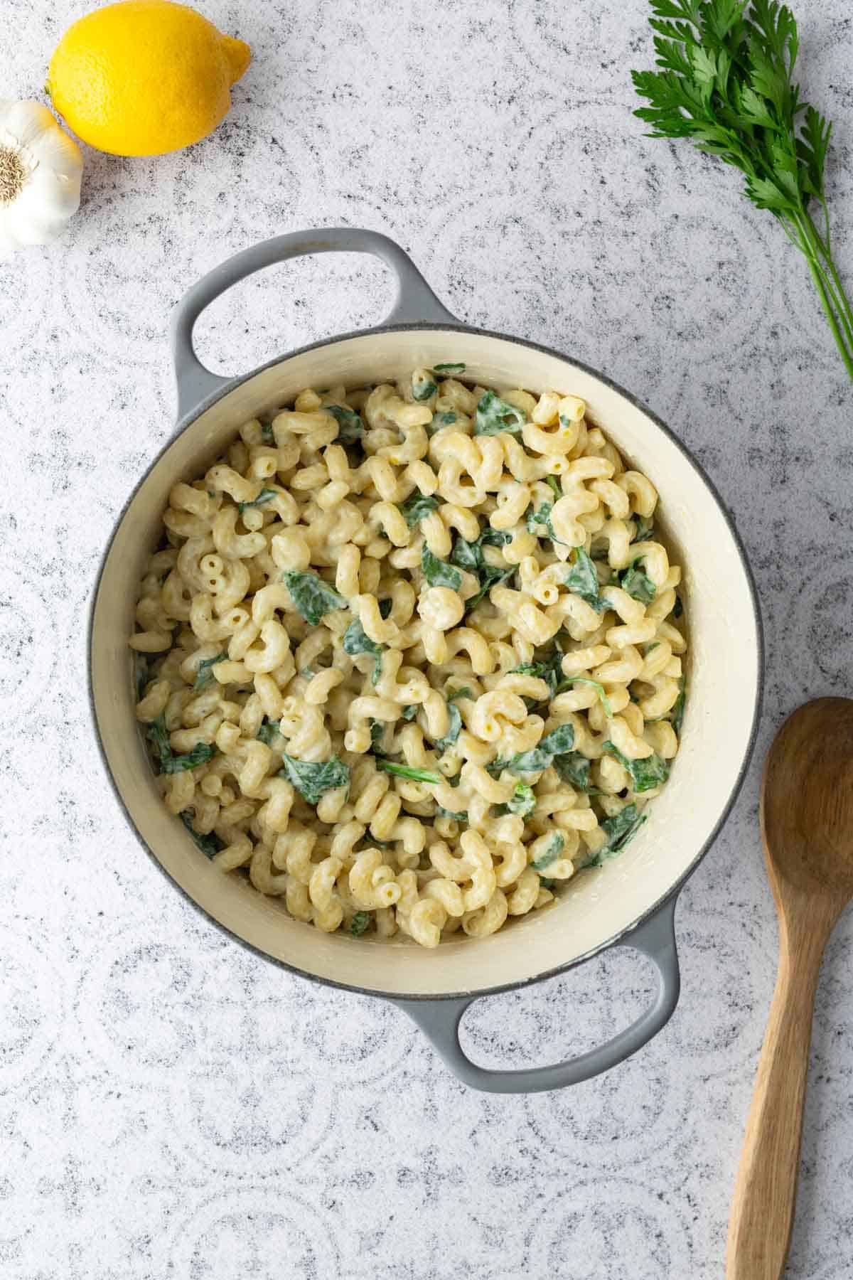 A pot of cooked cavatappi pasta mixed with spinach and a creamy sauce sits on a textured countertop next to a lemon, garlic, parsley, and a wooden spoon.