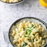 A bowl of creamy pasta garnished with parsley and shredded cheese, with a fork beside it. Another bowl, garlic, and a lemon are in the background.