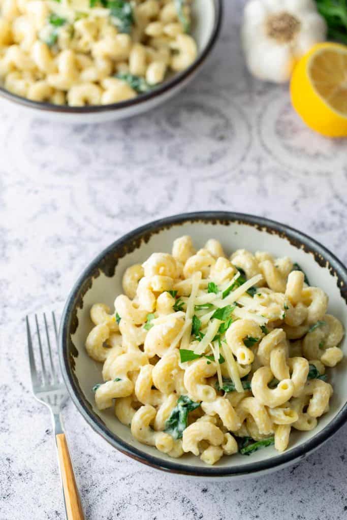 A bowl of creamy pasta garnished with parsley and shredded cheese, with a fork beside it. Another bowl, garlic, and a lemon are in the background.