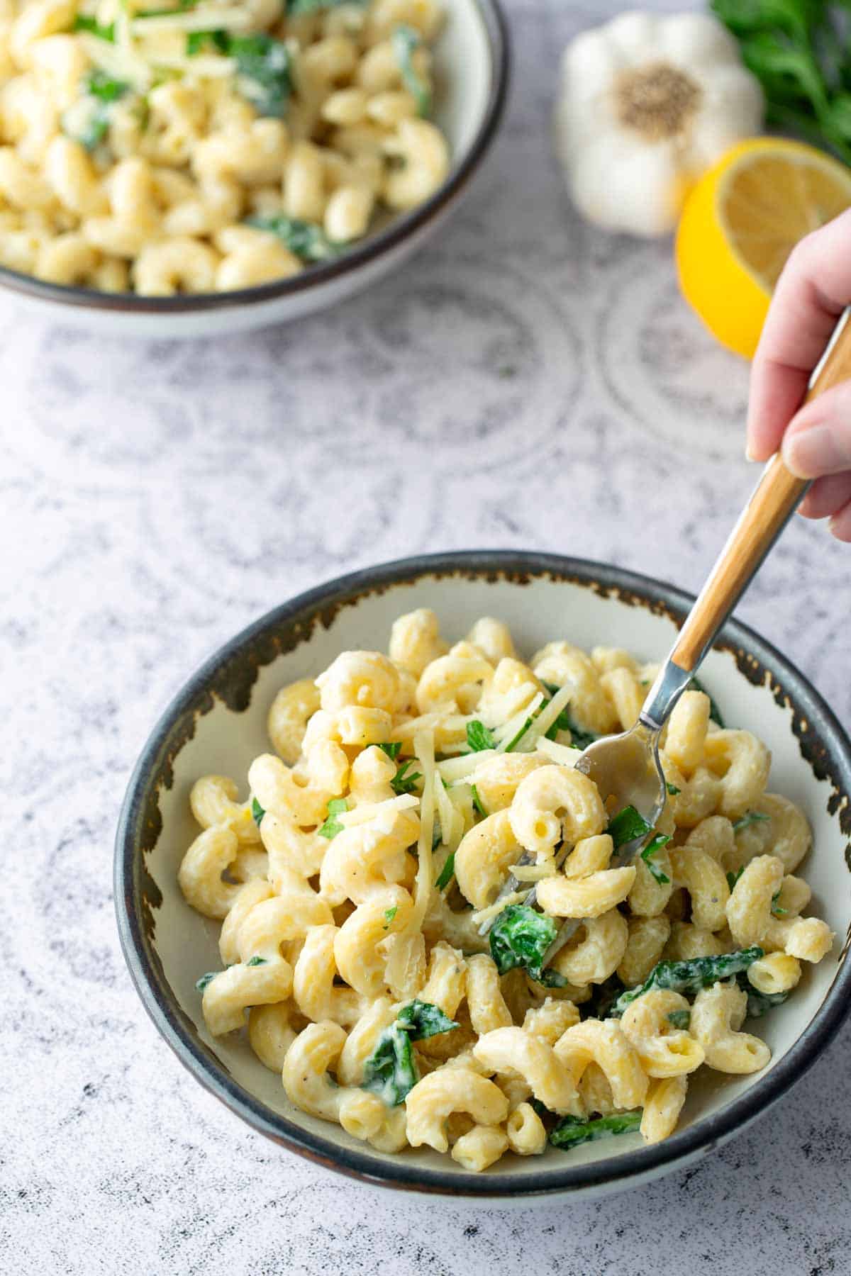 A bowl of creamy pasta with greens, with a hand holding a fork. Another bowl, garlic, and half a lemon are visible in the background.