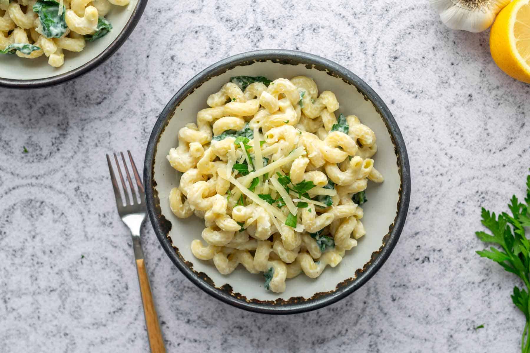 A bowl of creamy macaroni pasta garnished with fresh herbs and grated cheese, placed on a light textured surface beside a fork, garlic, lemon, and parsley.