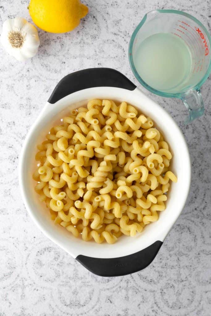 Cooked cavatappi pasta in a white colander, with a lemon, a garlic bulb, and a measuring cup of liquid on a light countertop.