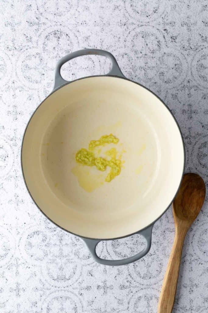 A small amount of minced garlic and oil in a large, empty, cream-colored pot beside a wooden spoon on a textured surface.