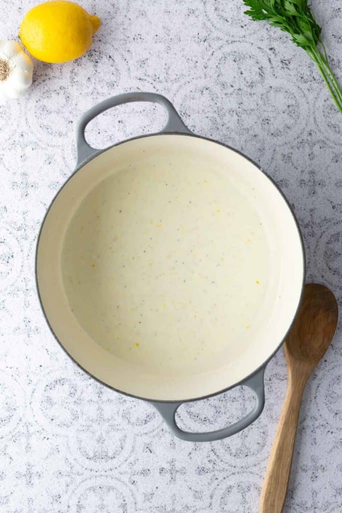 A pot of creamy white sauce sits on a patterned countertop next to a lemon, a garlic bulb, parsley, and a wooden spoon.