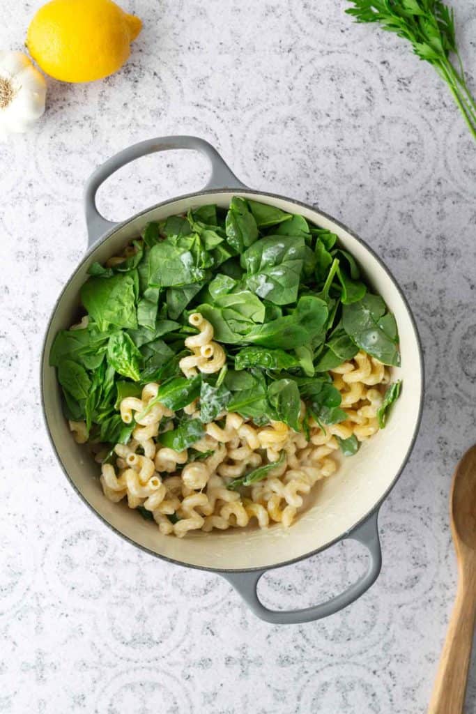 A pot of creamy pasta topped with fresh spinach leaves sits on a light countertop, with a lemon, garlic, and parsley nearby.