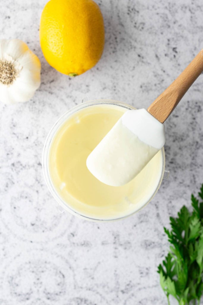 A bowl of creamy white sauce with a spatula, next to a lemon, garlic bulb, and fresh parsley on a light countertop.