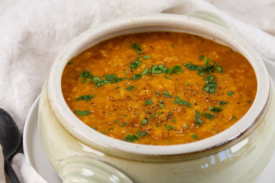 A ceramic bowl filled with orange lentil soup, garnished with chopped parsley and black pepper, placed on a light-colored cloth.
