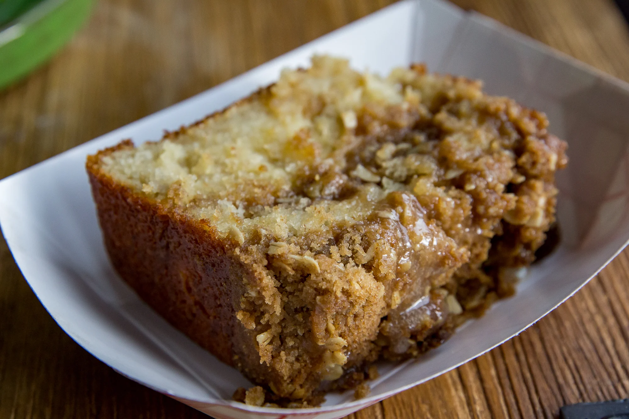 A slice of crumb cake with a brown sugar and oat topping on a white paper tray, placed on a wooden surface.