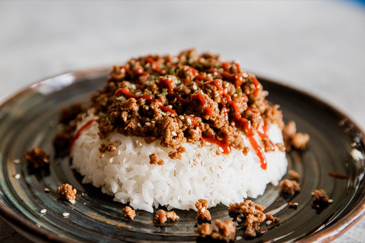 A serving of white rice topped with cooked ground meat, garnished with sauce and herbs, on a dark ceramic plate.