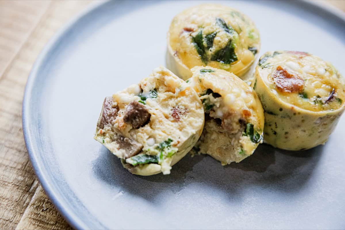 Three egg bites with spinach and mushrooms are arranged on a light gray plate, one of which is cut open to show the filling.