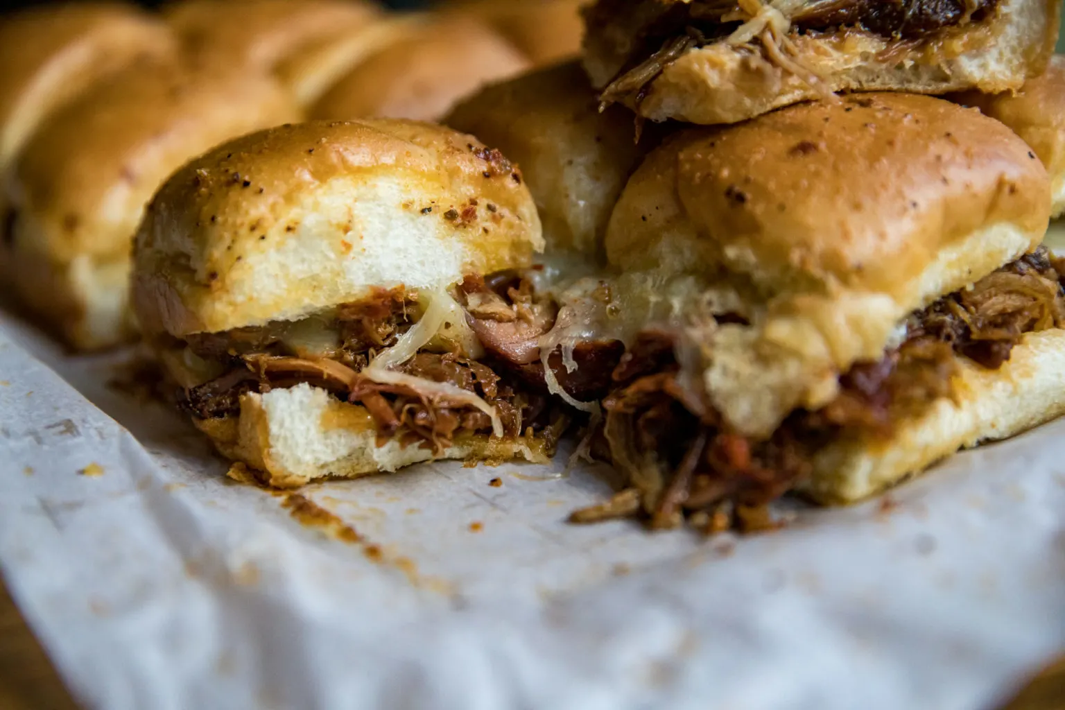Close-up of pulled pork sliders with melted cheese on soft, golden buns, arranged on a sheet of parchment paper.