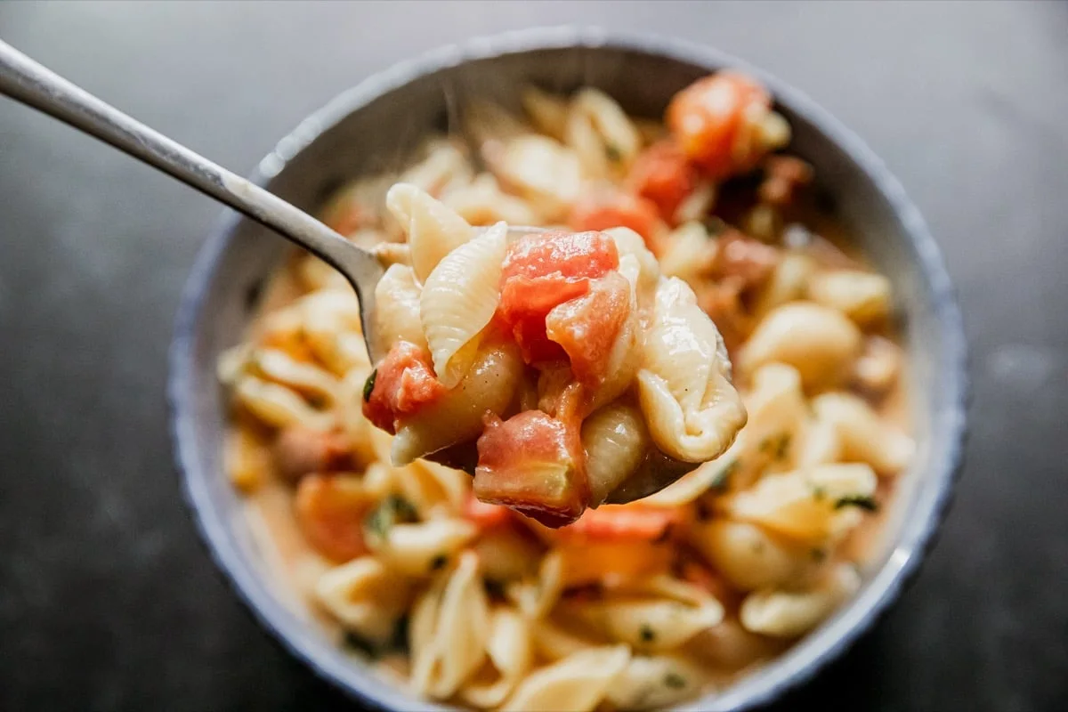A close-up of a spoon holding pasta shells mixed with diced tomatoes and sauce, with a bowl of the same pasta in the background.