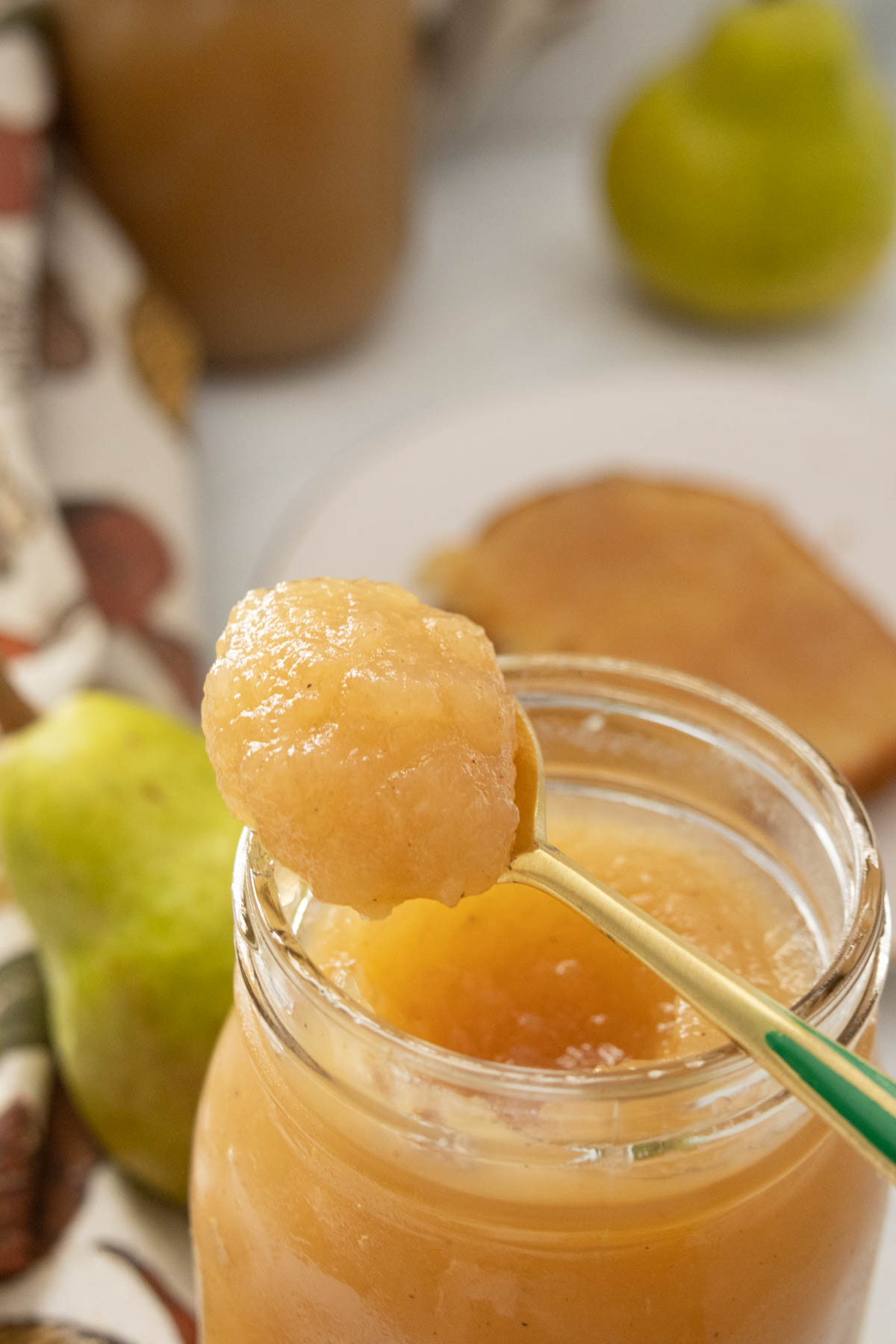 A spoon holding a portion of pear sauce above an open jar, with pears and a cloth in the background.