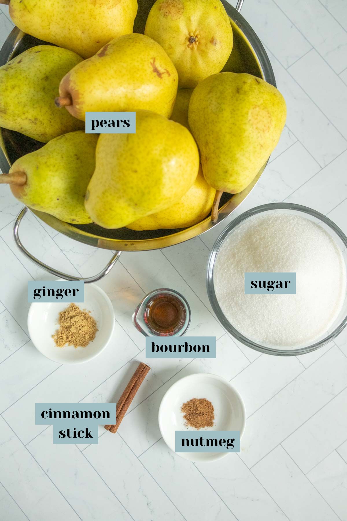 Overhead view of pears in a colander with bowls of sugar, ground ginger, ground nutmeg, a cinnamon stick, and a small glass of bourbon arranged on a white surface.
