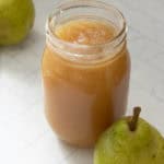 A glass jar filled with pear sauce sits on a white tiled surface next to a whole green pear.