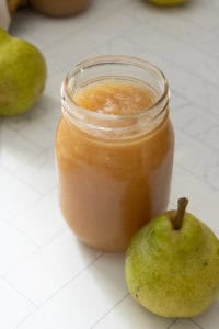 A glass jar filled with pear sauce sits on a white tiled surface next to a whole green pear.