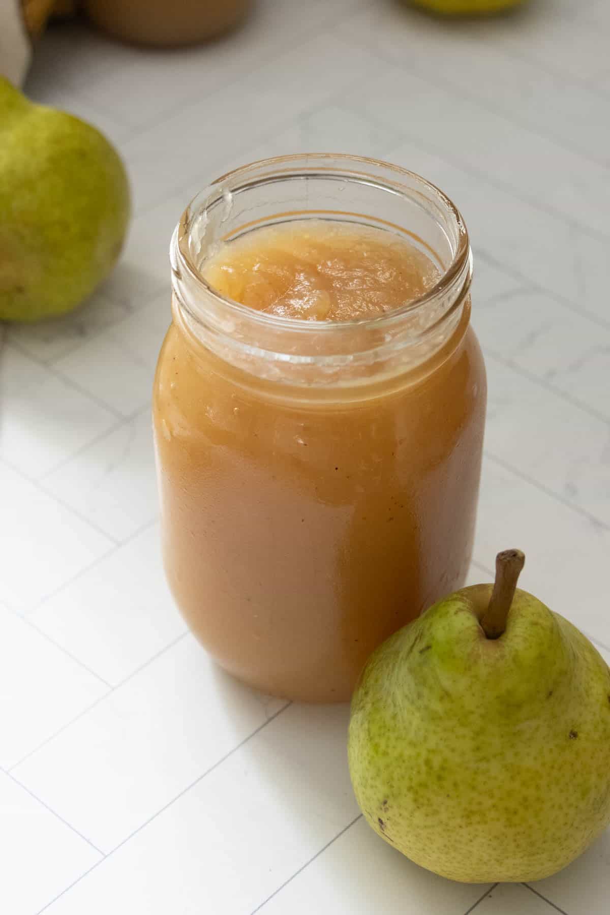 A glass jar filled with pear sauce sits on a white tiled surface next to a whole green pear.