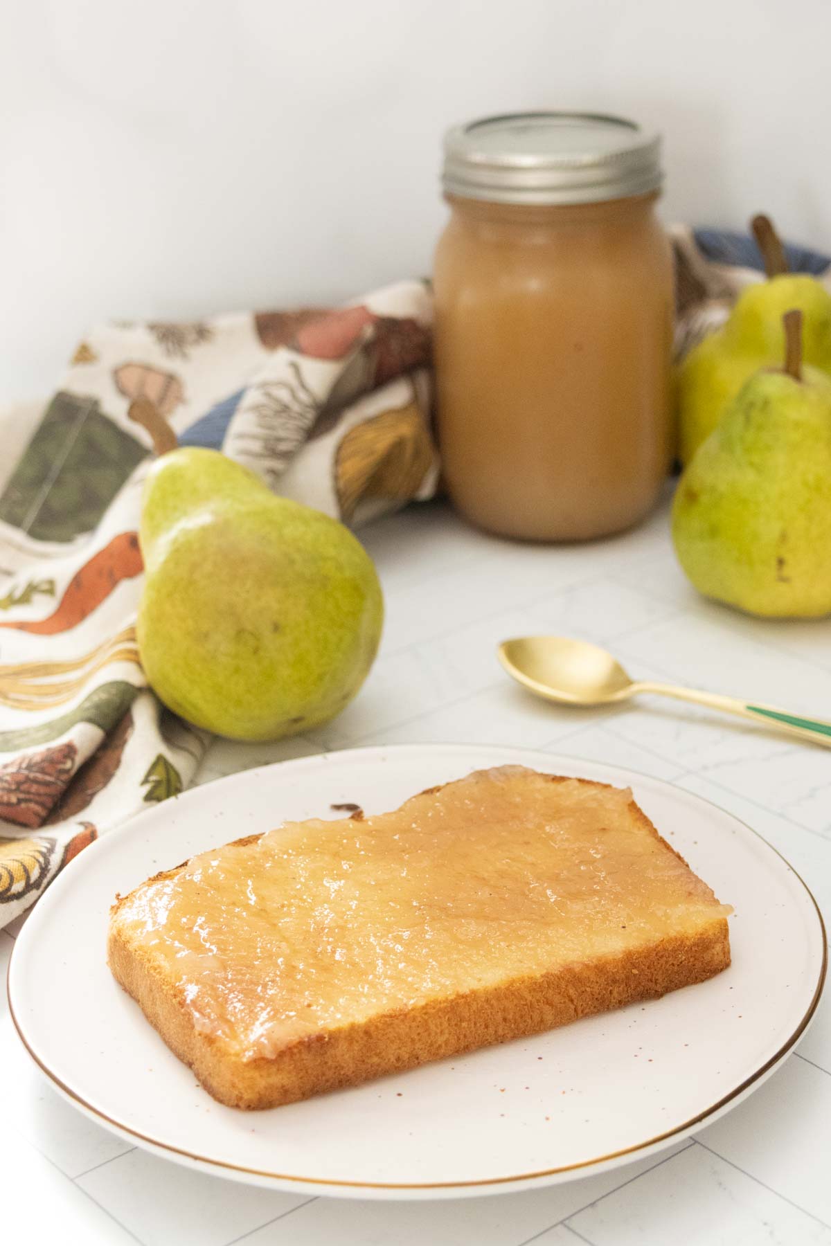A slice of bread with pear jam on a plate, with a jar of pear jam, two fresh pears, a gold spoon, and a patterned cloth in the background.