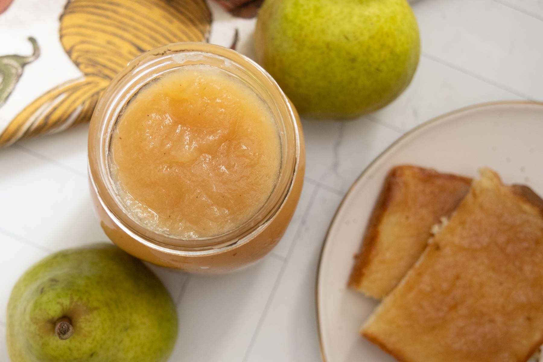 An open jar of pear sauce is on a table with two fresh pears and a plate holding slices of bread.