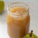 A mason jar filled with spiced bourbon pear butter sits on a white surface next to a fresh pear.