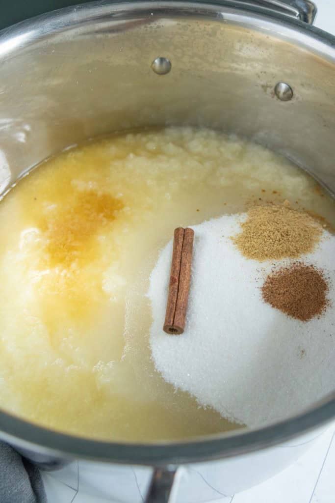 A stainless steel pot containing applesauce, granulated sugar, a cinnamon stick, ground spices, and vanilla extract, ready to be mixed.