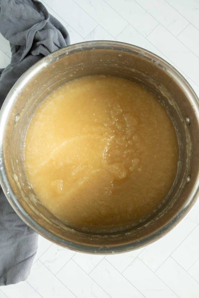 A pot filled with smooth, light brown applesauce sits on a white tiled surface next to a gray cloth.