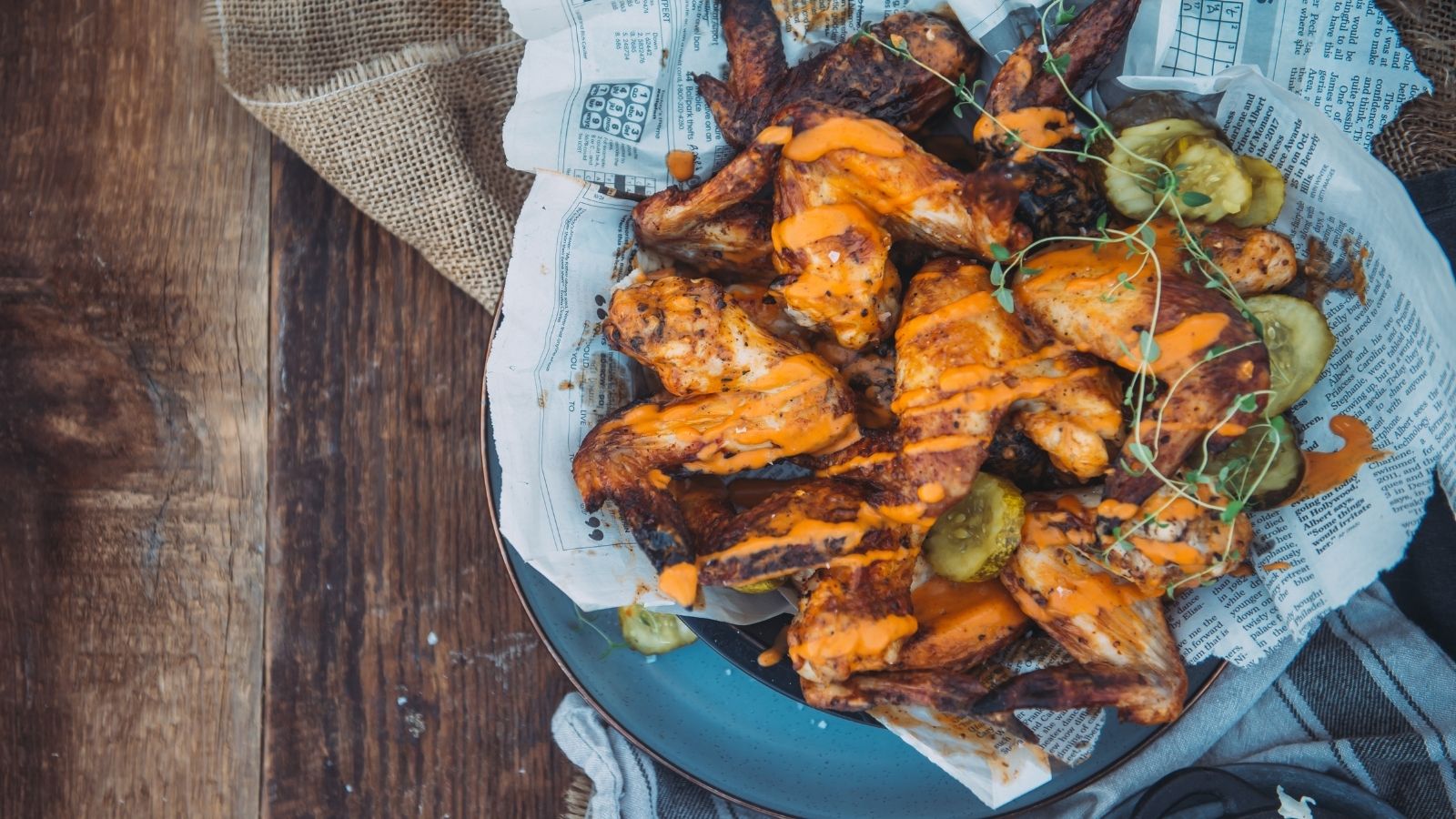A plate of grilled chicken wings with orange sauce, garnished with microgreens and pickles, served on newspaper print paper.